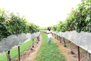 Kim standing in a vineyard with a white dress and jeans on a bright and sunny day.